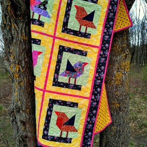 Op de afbeelding: Een gele quilt met een zwarte, roze en groene rand. De quilt heeft vier kleurrijke vogelontwerpen in het midden.
