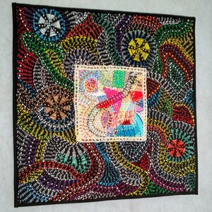 May include: A colorful abstract quilt with a central square featuring a smaller, more detailed abstract design. The quilt is made of various fabrics and threads, creating a textured and intricate pattern.