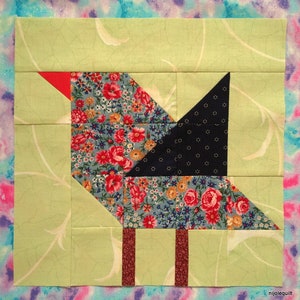 Op de afbeelding: Een patchwork quiltblok met een vogelontwerp. De vogel is gemaakt van verschillende stoffen, waaronder een bloemenprint, een donkerblauwe stof met witte stippen en een rode stof. De vogel staat op twee bruine poten.