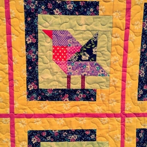 Op de afbeelding: Een quiltblok met een gele achtergrond en roze, paarse en zwarte stof. Het blok heeft een vogelontwerp met een rode snavel en een groene stengel.