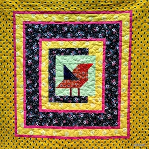 Op de afbeelding: Een kleurrijk quilt met een rood, oranje en blauw vogeltje in het midden. De quilt is omgeven door een gele, roze en zwarte rand.