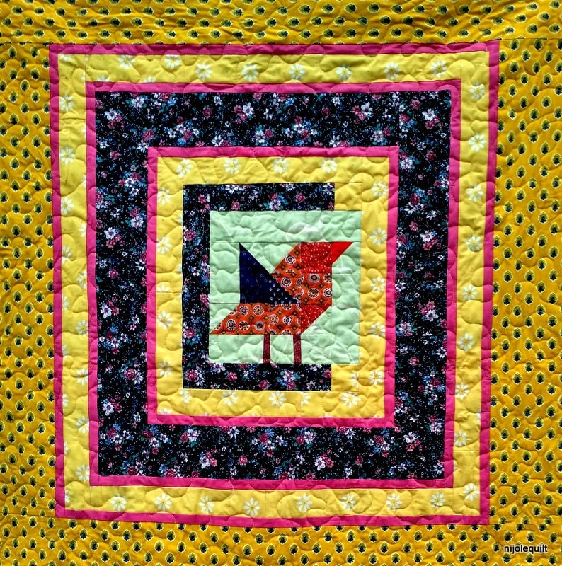 Op de afbeelding: Een kleurrijk quilt met een rood, oranje en blauw vogeltje in het midden. De quilt is omgeven door een gele, roze en zwarte rand.