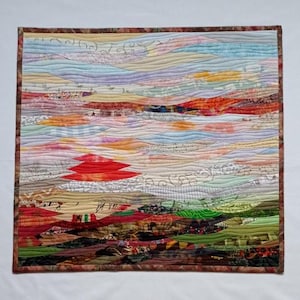 Op de afbeelding: Een kleurrijk abstract quilt met een rood bergketen in het midden. De quilt is gemaakt van verschillende stoffen en texturen, waardoor een gelaagd en textuurrijk effect ontstaat. De quilt is omlijst met een bruine rand.