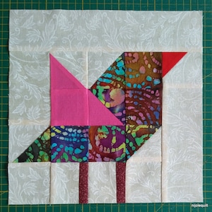 Op de afbeelding: Een patchwork quiltblok met een vogelontwerp in roze, rood, bruin, blauw en groene stof. De vogel heeft een lange snavel en drie poten. De achtergrond is een witte stof met een bloemenpatroon.