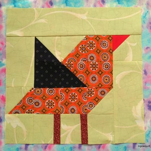 Op de afbeelding: Een patchwork quiltblok met een gestileerd vogelontwerp. De vogel is gemaakt van oranje, zwart en rood stof met een groene achtergrond. De vogel heeft een lange, dunne snavel en een klein, rond hoofd. Het lichaam van de vogel is gemaakt van een gestreept stof met kleine cirkels en vierkanten. De vleugels van de vogel zijn gemaakt van een effen zwart stof. De poten van de vogel zijn gemaakt van een effen rood stof.
