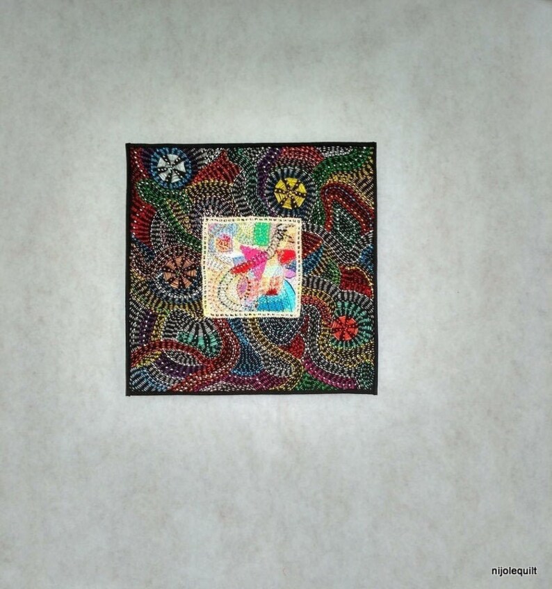 May include: A colorful square quilt block with a black border. The block is made of various fabrics and colors, including red, green, blue, yellow, and white. The center of the block features a smaller square with a geometric design.