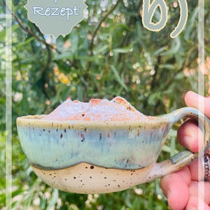 Könnte beinhalten: Ein Keramikbecher mit cremefarbener, gesprenkelter Basis und hellgrünem glasiertem Rand, der ein mit Puderzucker bestäubtes Gebäck enthält. Der Text "+ Rezept" und "b)" ist im oberen Teil des Bildes zu sehen.