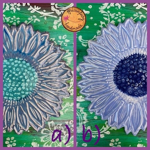 May include: Two ceramic sunflower designs, one with a teal center and the other with a dark blue center, are displayed side-by-side. The petals are a light blue-gray color. The background is green with white floral patterns. The image includes the text "a)" and "b)".