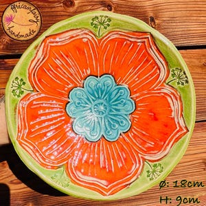 Könnte beinhalten: Handgefertigte Keramikplatte mit einem leuchtend orangefarbenen Blumenmuster und einem türkisfarbenen Zentrum. Der Rand ist grün mit floralen Akzenten. Durchmesser 18cm, Höhe 9cm. Von Africanfairy handmade.