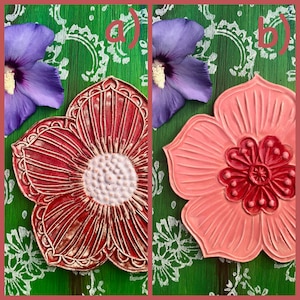 May include: Two ceramic flower-shaped coasters. One is red with a white center and detailed lines, the other is pink with a darker red center. The background is green with white floral patterns and purple flowers.