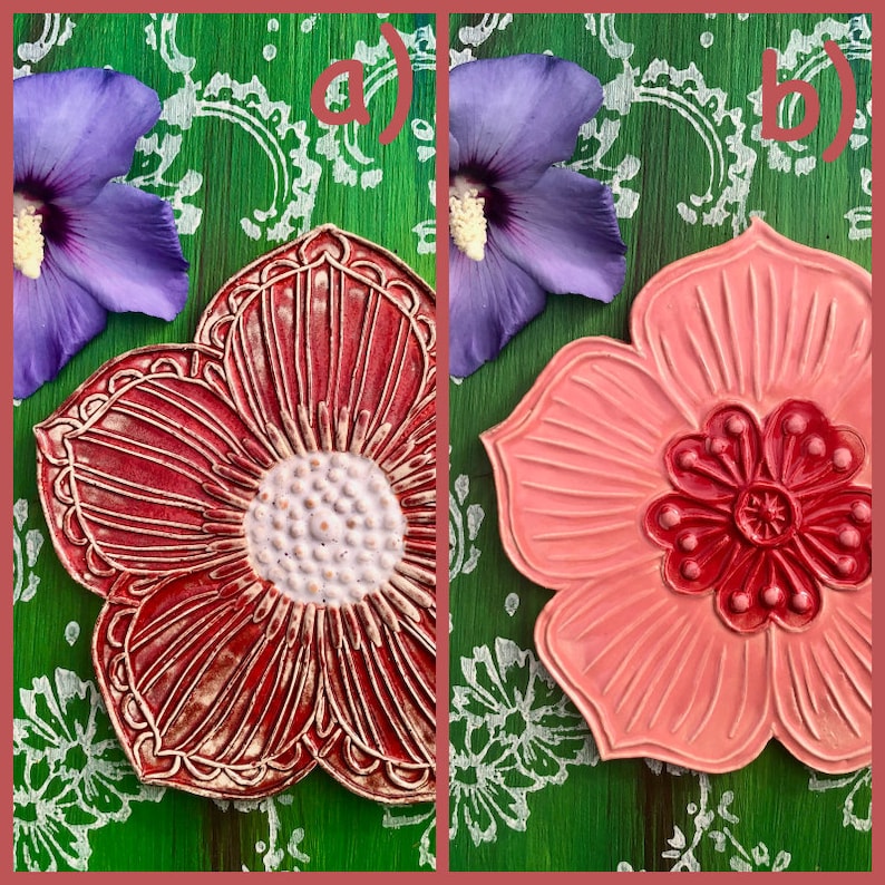 May include: Two ceramic flower-shaped coasters. One is red with a white center and detailed lines, the other is pink with a darker red center. The background is green with white floral patterns and purple flowers.