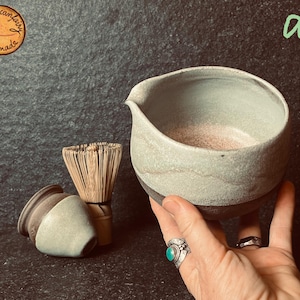 Könnte beinhalten: Handgefertigte Matcha-Schale aus Keramik mit Ausguss, gehalten von einer Hand. Die Schale ist hellgrün mit einem dunkelbraunen Boden. Ein Bambusbesen und ein kleiner Keramikbehälter sind ebenfalls zu sehen. Africanfairy Handmade Logo.