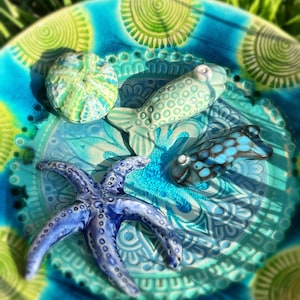 Könnte beinhalten: Eine runde Keramikschale mit blau-grünem Spiralmuster, die vier dekorative Figuren zum Thema Meer enthält: einen hellgrünen Seeigel, einen blassgrünen Fisch, einen dunkelblauen Seestern und einen kleinen Fisch mit blauen und schwarzen kreisförmigen Markierungen.
