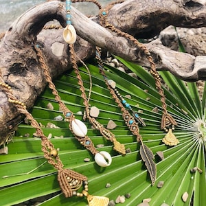 Op de afbeelding: Vijf macramé kettingen met schelp- en metalen bedels. De kettingen zijn bruin en hebben turquoise kralen. De kettingen worden getoond op een groen palmblad.