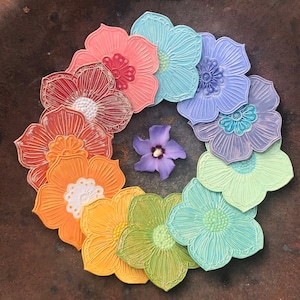 May include: A circular arrangement of colorful, flower-shaped coasters in a rainbow of hues, including red, orange, yellow, green, blue, and purple. Each coaster has a detailed floral design. A single purple flower is in the center.