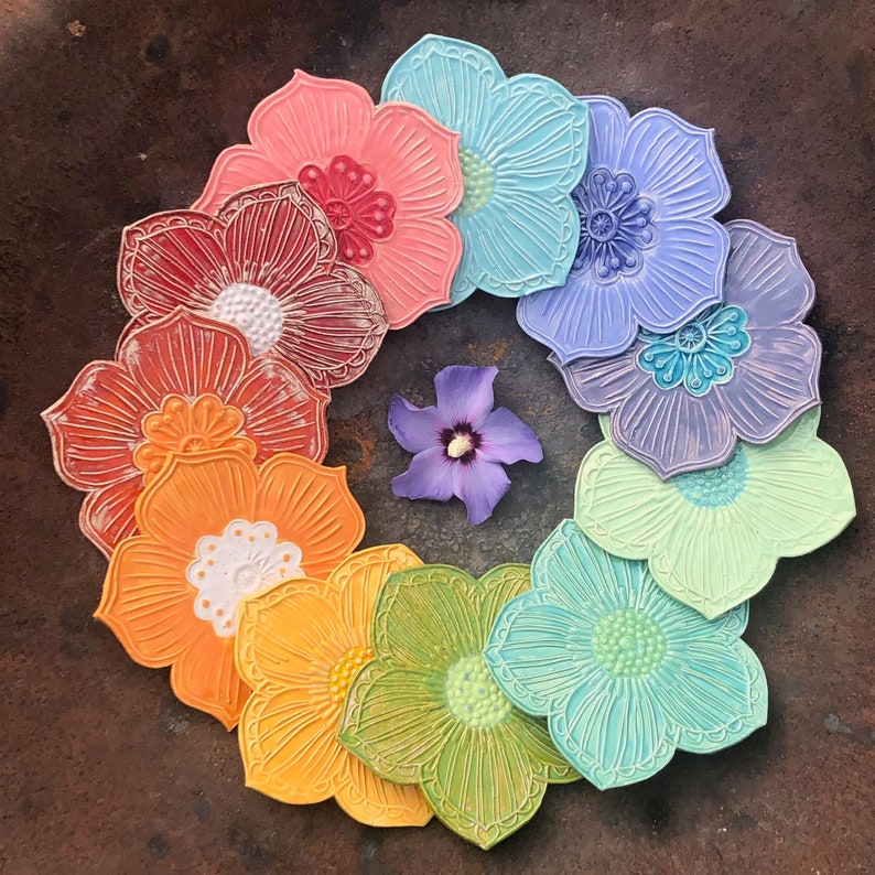 May include: A circular arrangement of colorful, flower-shaped coasters in a rainbow of hues, including red, orange, yellow, green, blue, and purple. Each coaster has a detailed floral design. A single purple flower is in the center.