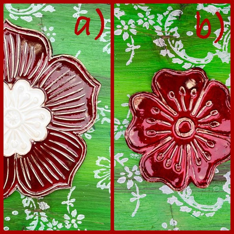 May include: Two ceramic flower ornaments. One has a white center and burgundy petals, the other is all burgundy. Both are on a green background with white floral patterns. The ornaments are framed in red.