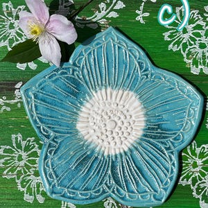 May include: A turquoise ceramic flower-shaped dish with a white center and detailed carved lines. A light pink flower with a yellow center and green leaves sits near the dish. The letter "c)" is in the upper right corner.