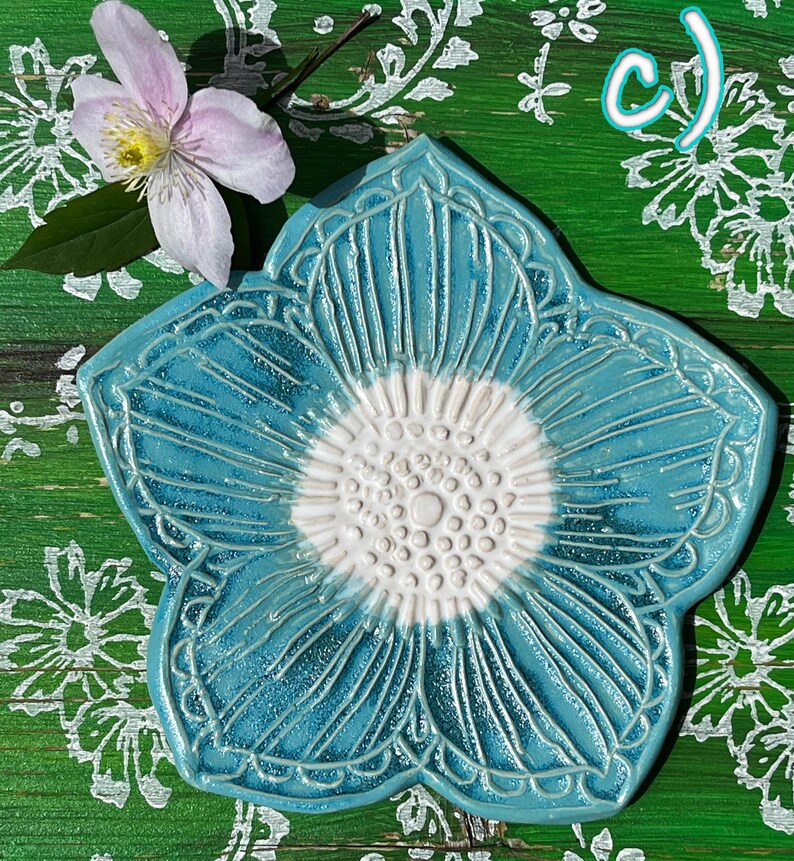 May include: A turquoise ceramic flower-shaped dish with a white center and detailed carved lines. A light pink flower with a yellow center and green leaves sits near the dish. The letter "c)" is in the upper right corner.