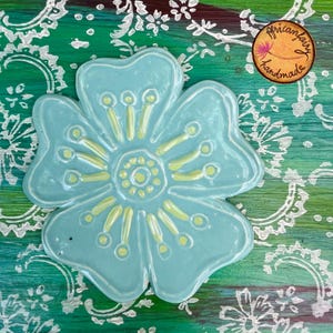 May include: A light blue ceramic flower with yellow accents. The handmade floral design is on a green and white patterned background. The image includes a circular sticker with the text "Africanfairy handmade".
