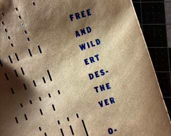 Könnte beinhalten: Ein beigefarbenes Blatt Papier mit dunkelblauem Aufdruck, der FREE AND WILD ERT DES- THE VER 0. VOICE lautet. Das Papier weist links neben dem Text eine Reihe von vertikalen, rechteckigen Stanzungen in Spalten auf.