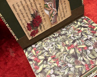 May include: Open journal with decorative pages. The interior features a botanical print with red berries and green leaves. The journal's cover has a dark green pocket with text and a floral embellishment. The journal is open on a red surface.