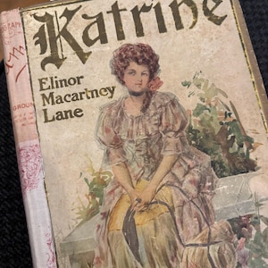 May include: A vintage hardback book titled Katrine by Elinor Macartney Lane. The cover features a colour illustration of a person with dark hair wearing a ruffled dress, sitting outdoors. The title is printed in a bold, dark, decorative font.