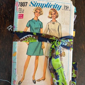 Pode incluir: Um diário feito à mão com uma capa de molde de costura vintage Simplicity 7807 ilustrando duas figuras em vestidos. A capa é amarrada com uma fita de tecido estampada em azul escuro, verde e marrom e inclui cantoneiras metálicas.