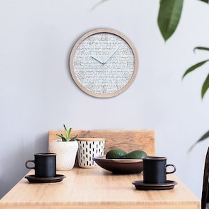 Op de afbeelding: Een ronde, witte klok met zwarte Romeinse cijfers op een lichte houten achtergrond. De klok hangt aan een lichtgrijze muur. De klok bevindt zich in een eetkamer met een houten tafel, twee stoelen en een potplant.