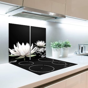 Puede incluir: Salpicadero de cocina negro y blanco con un diseño floral con nenúfares blancos sobre un fondo negro. El salpicadero está instalado detrás de una placa de cocina eléctrica negra con cuatro quemadores redondos.