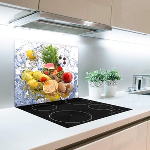 Puede incluir: Un protector de salpicaduras de cocina con un diseño de frutas coloridas. El protector de salpicaduras presenta una variedad de frutas, incluyendo piñas, plátanos, limones, sandía, fresas y kiwi, todo rodeado de gotas de agua. El protector de salpicaduras está instalado detrás de una estufa eléctrica negra.