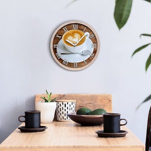 Puede incluir: Un reloj redondo de madera con una taza de café blanca con arte latte sobre un platillo blanco. La esfera del reloj tiene números romanos y el fondo es un patrón de veta de madera.