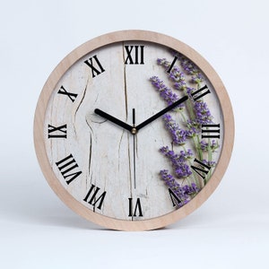 Puede incluir: Un reloj redondo de madera con números romanos y un diseño floral de lavanda. La esfera del reloj es blanca con una textura de madera desgastada.