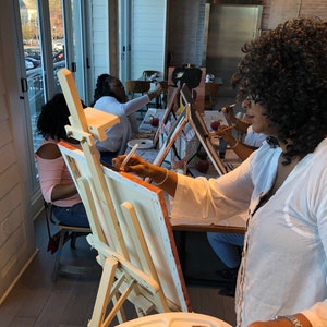 May include: A group of people painting at easels in a well-lit room. Each person has a canvas and a palette with various paint colors. The scene suggests a painting class or social event focused on art.