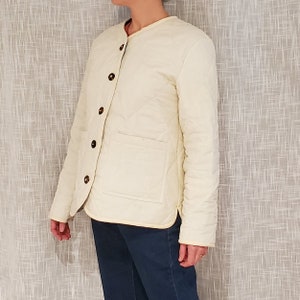 May include: A cream-colored quilted jacket with a stand-up collar and button closure. The jacket has a patch pocket on the left side and a diamond-shaped quilting pattern.