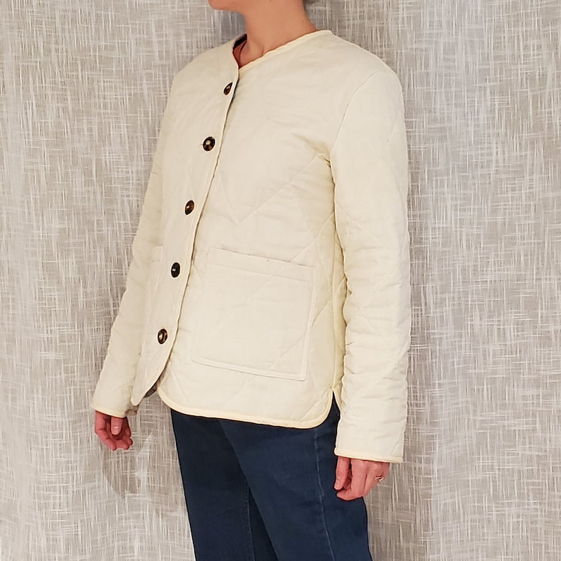 May include: A cream-colored quilted jacket with a stand-up collar and button closure. The jacket has a patch pocket on the left side and a diamond-shaped quilting pattern.