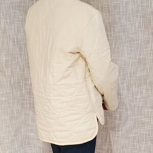 May include: A white quilted jacket with a long sleeve and a diamond pattern. The jacket has a stand-up collar and a zipper closure.