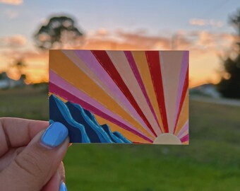 May include: A small, rectangular sticker featuring a colorful sunset design. The artwork depicts a mountain range in shades of blue, with a vibrant sunburst in hues of orange, pink, and red. The sticker is held against a blurred background of a sunset.