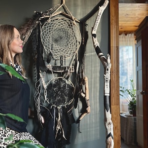 Puede incluir: Un atrapasueños grande, hecho a mano, con un diseño de red tejida, plumas y cuentas. El atrapasueños es principalmente negro y crema, con detalles de madera natural. También se ve una rama decorativa de madera con patrones geométricos en blanco y negro.