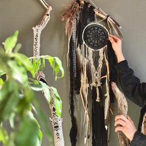 Puede incluir: Un atrapasueños hecho a mano con un diseño de red negro, plumas y detalles de macramé. El atrapasueños está suspendido de una rama de madera, adornado con encaje negro y hierbas secas. También se ve una rama de madera decorativa con diseños pintados.