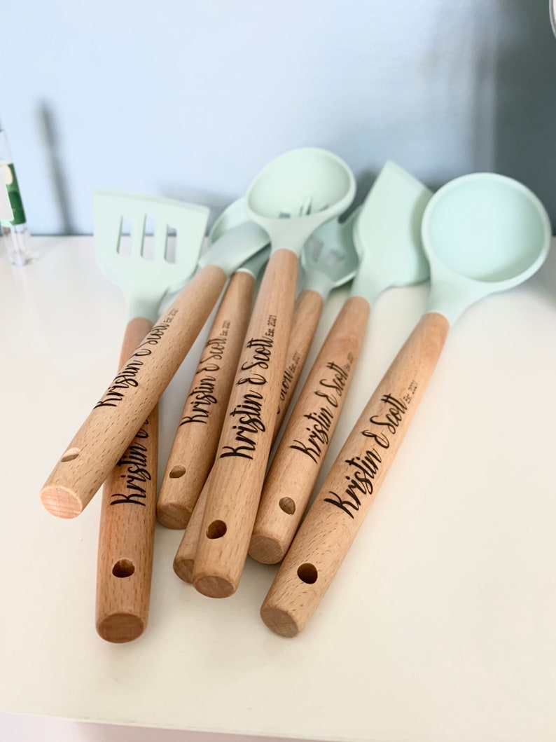 May include: A set of six wooden handled kitchen utensils with light blue silicone heads. The handles are engraved with the names "Kristin & Scott" and the date "October 22, 2022".