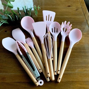 May include: A set of pink silicone kitchen utensils with wooden handles. The set includes a spatula, spoon, ladle, whisk, tongs, and a pasta server.