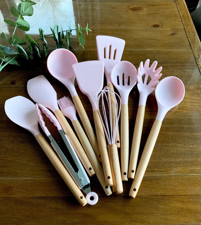 May include: A set of pink silicone kitchen utensils with wooden handles. The set includes a spatula, spoon, ladle, whisk, tongs, and a pasta server.