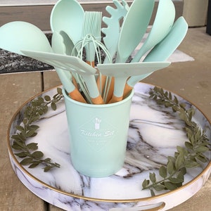 May include: A set of light green silicone kitchen utensils with wooden handles in a light green ceramic container with the text "Kitchen Set Food & Drinks" on a white marble tray with green leaves.