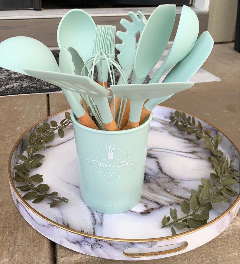 May include: A set of light green silicone kitchen utensils with wooden handles in a light green ceramic container with the text "Kitchen Set Food & Drinks" on a white marble tray with green leaves.