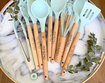 Puede incluir: Un juego de nueve utensilios de cocina de silicona azul claro con mangos de madera. Los mangos están grabados con el texto "Chef Reefski".