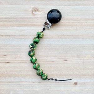 Peut inclure: Un hochet pour bébé en silicone vert et noir camouflage avec un clip en bois noir. Le hochet est enfilé sur un cordon noir.