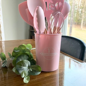May include: A pink kitchen utensil holder with a set of pink silicone kitchen utensils, including a spatula, spoon, whisk, and tongs. The holder has a white logo that says "Kitchen & Dining".