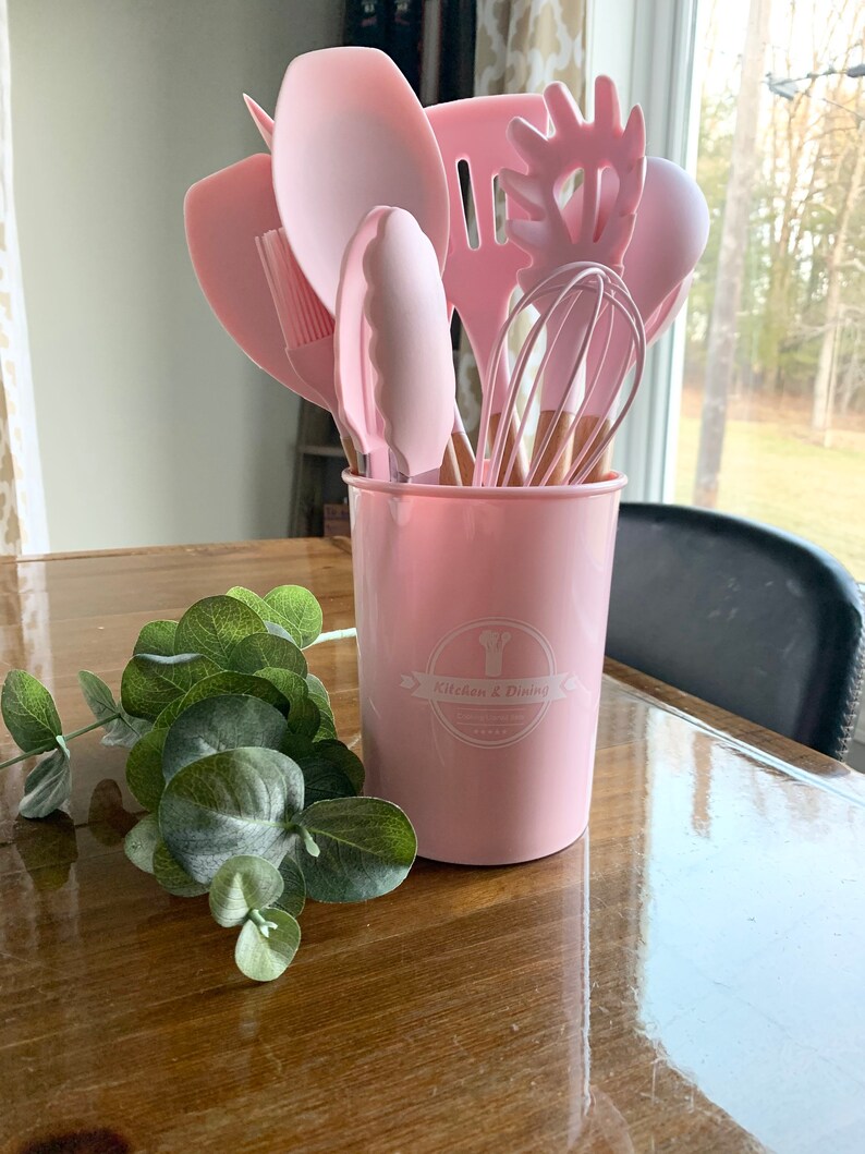 May include: A pink kitchen utensil holder with a set of pink silicone kitchen utensils, including a spatula, spoon, whisk, and tongs. The holder has a white logo that says "Kitchen & Dining".