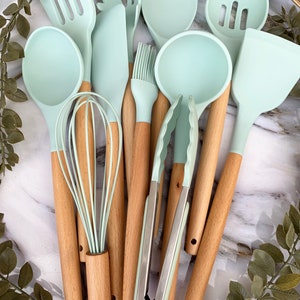May include: A set of ten kitchen utensils with light blue silicone heads and wooden handles. The utensils include a spatula, a spoon, a whisk, a ladle, a slotted spoon, a basting brush, a set of tongs, and a spatula with a scalloped edge.